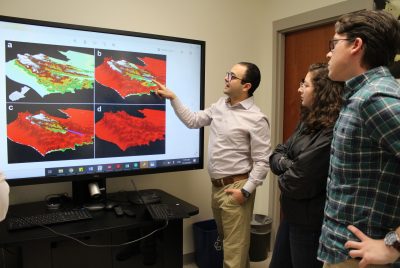 Two men and a woman stand looking at a large computer screen displaying an array of four maps. One of the men points to one of the images.