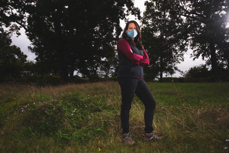 A woman wearing a face covering standing with her arms crossed in a grassy area with trees in the background