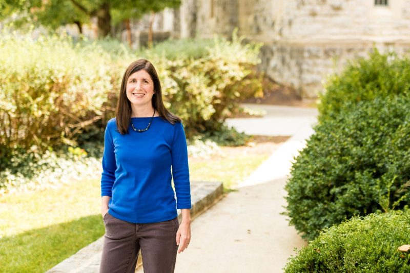 A woman with shoulder-length brown hair and wearing a blue long sleeve shirt, poses near some green bushes on a bright, sunny day.